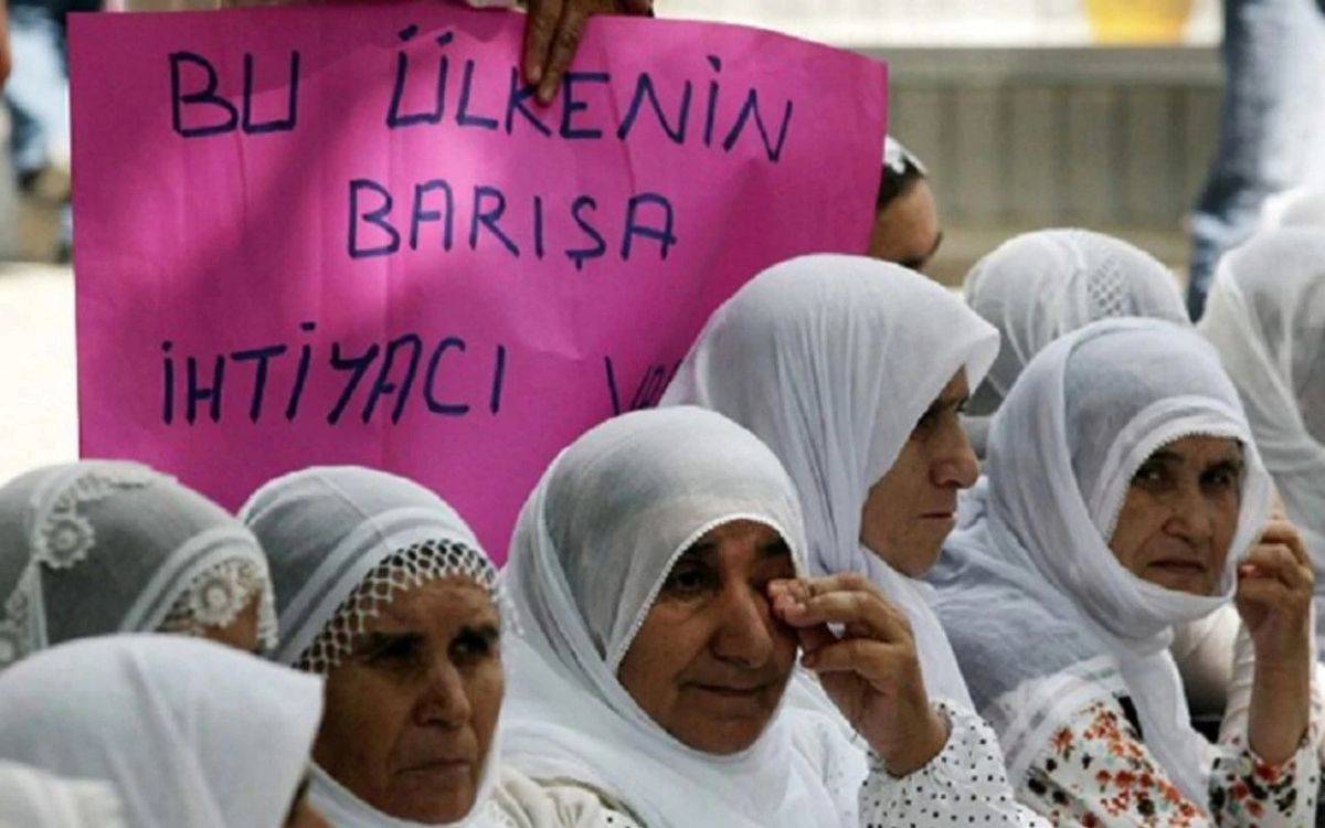 Bu fotoğrafta, yaşları ileri olan kadınlar beyaz başörtüleriyle bir arada oturuyorlar. Kadınların çoğu hüzünlü bir şekilde yere ya da bir noktaya bakıyor; bazıları ise gözyaşlarını siliyor. Fotoğrafın arka planında, bir kişinin tuttuğu pembe bir pankart öne çıkıyor. Pankartta büyük harflerle el yazısıyla "Bu ülkenin barışa ihtiyacı var" ifadesi yer alıyor. Kadınların kıyafetleri sade ve geleneksel, başörtüleri beyaz ve işlemeli. Fotoğraf, barış talebinde bulunan kadınların duygusal ve kararlı duruşunu simgeliyor, ülkenin barışa duyduğu ihtiyacı güçlü bir şekilde vurguluyor.