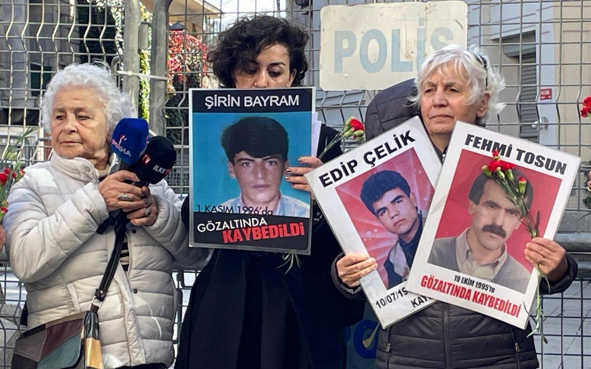 Fotoğrafta, yaşları ilerlemiş kadınlar ellerinde kayıp yakınlarının fotoğraflarını taşıyarak bir protesto eyleminde bulunuyor. Polis bariyerlerinin önünde gerçekleştirilen bu eylemde kadınlar, kaybedilen yakınlarının adalet arayışını temsil eden afişler tutuyor. Afişlerde üç kayıp insanın fotoğrafları ve isimleri yer alıyor: Şirin Bayram, Edip Çelik ve Fehmi Tosun. Her bir fotoğrafın altında "Gözaltında Kaybedildi" ifadesi bulunuyor. Kadınların yüzünde ciddi bir ifade var ve ellerinde taşıdıkları karanfiller, bu kayıpları anmak amacıyla kullanılmış. Fotoğrafın arka planında polis bariyerleri dikkat çekiyor, bu da eylemin güvenlik önlemleri altında gerçekleştirildiğini gösteriyor.