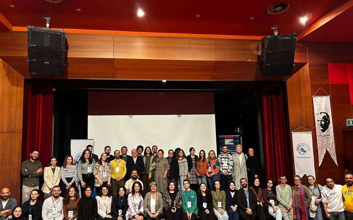 Bu fotoğraf, bir konferans ya da etkinlik sonrasında topluca poz veren bir grup katılımcıyı göstermektedir. Katılımcılar, sahnede sıralanarak grup halinde poz vermiş. Arkada geniş bir perde, sağ ve sol taraflarda etkinliğin destekleyicilerinin veya organizatörlerin logoları yer alıyor. Sahne tasarımı ahşap ve kırmızı detaylarla çevrili olup, resmi bir etkinlik atmosferi oluşturuyor.  Katılımcılar arasında farklı yaş gruplarından insanlar bulunuyor; bazıları boyunlarında etkinlik kimlik kartları taşımakta. Çoğu kişi kameraya bakarak gülümsüyor, etkinliğin sıcak ve samimi bir ortamda geçtiği izlenimi veriyor. Mekanın üst kısmında ahşap kaplama ve büyük hoparlörler dikkat çekiyor. Fotoğraf, bir araya gelmiş bu grup insanın dayanışma ve birliktelik ruhunu yansıtarak etkinliğin başarıyla tamamlandığını simgeliyor.