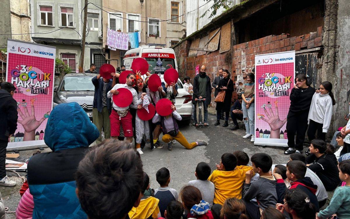 Fotoğrafta, İstanbul Büyükşehir Belediyesi tarafından düzenlenen "3. Çocuk Hakları Festivali" etkinliğinden bir sahne görülüyor. Dar bir sokakta gerçekleşen etkinlikte, palyaço kostümleri giymiş bir grup sanatçı, kırmızı renkli yuvarlak aksesuarlar kullanarak çocuklara bir gösteri sunuyor. Çevredeki çocuklar dikkatle ve keyifle gösteriyi izliyor. Sahnenin yanında, üzerinde "3. Çocuk Hakları Festivali" yazılı büyük bir afiş bulunuyor. Afişte etkinliğin 11-20 Kasım 2023 tarihleri arasında gerçekleştirildiği belirtilmiş.