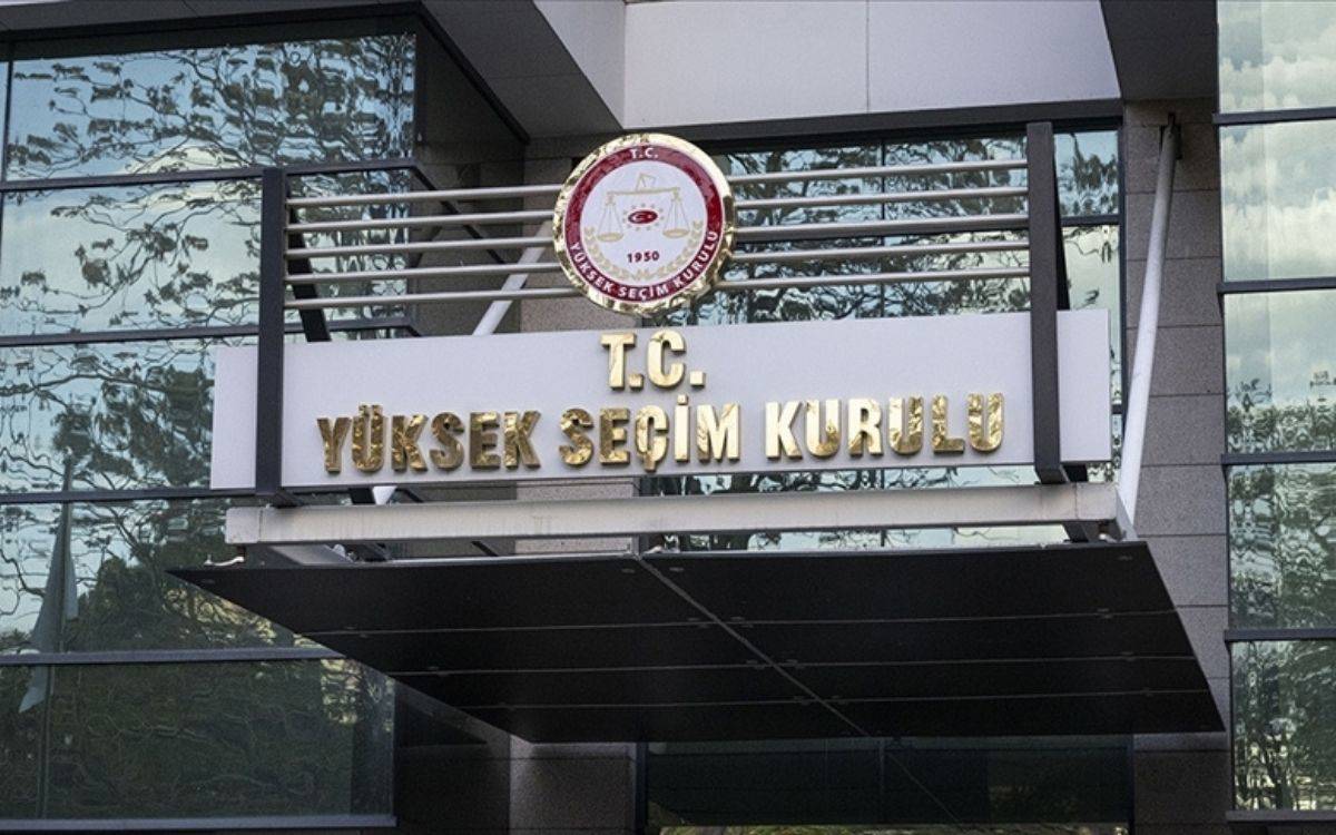 Bu görselde, Türkiye Cumhuriyeti Yüksek Seçim Kurulu'nun (YSK) bina girişine ait bir görüntü yer almaktadır. Girişte, YSK'nın adını belirten bir tabela dikkat çekmektedir. Tabelada "T.C. Yüksek Seçim Kurulu" ifadesi altın sarısı büyük harflerle yazılmıştır. Tabelanın üst kısmında YSK'nın logosu bulunmakta, logoda Türkiye Cumhuriyeti'ne ait kırmızı-beyaz renkte bir amblem yer almaktadır. Binanın modern bir mimariye sahip olduğu görülmektedir; büyük cam paneller ve gri renkli metal detaylar dikkat çekmektedir. Tabelanın etrafında ağaçların ve doğanın yansımasını gösteren cam yüzeyler bulunmaktadır.