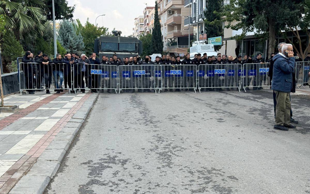 Bu fotoğrafta, Türkiye’de bir şehirde polis ekipleri, yolun ortasına yerleştirilen metal bariyerlerin arkasında sıralanmış şekilde görülüyor. Polis memurları, yoğun bir güvenlik önlemi alarak yolu kapatmış durumda. Bariyerlerin arkasında çok sayıda polis memuru topluca bekliyor. Arkada, bir polis aracı ya da zırhlı araç dikkat çekiyor. Ön planda ise iki sivil erkek, polis bariyerlerine yakın bir noktada duruyor; bir tanesi cep telefonuyla konuşuyor. Çevrede ise ağaçlar ve apartmanlar gözlemleniyor. Görüntü, güvenlik önlemlerinin yüksek olduğu bir durumu yansıtıyor.