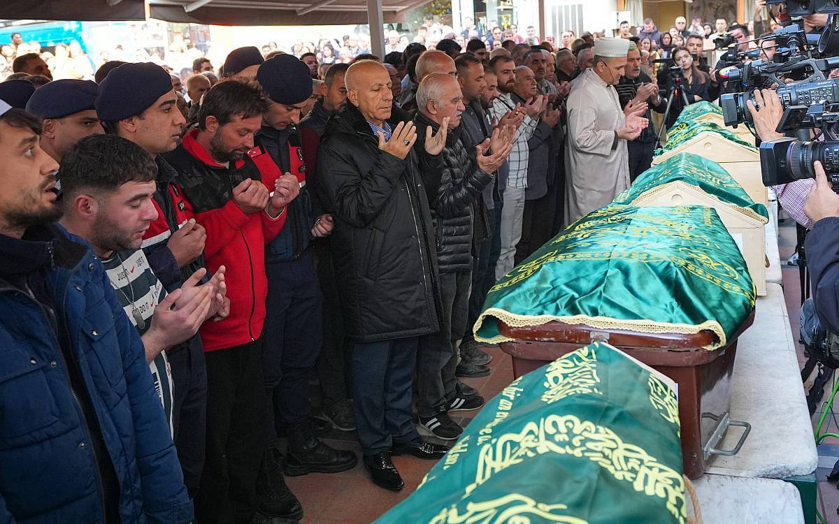 Bu fotoğrafta, bir cenaze töreni sırasında kalabalık bir grup insan dua ederken görülüyor. Yeşil örtülerle kaplanmış tabutlar yan yana dizilmiş, tabutların üzerinde Arapça yazılar yer alıyor. Fotoğrafta, ellerini dua pozisyonunda açmış, üzüntü içindeki insanlar cenazeye saygılarını gösteriyor. Kalabalık içinde jandarma ve sağlık görevlileri gibi resmi kıyafetli kişilerin yanı sıra, sivil kıyafetli vatandaşlar da bulunuyor. Arka planda, imam dua yönetiyor. Medya mensupları ise cenazeyi ve törene katılanları kayda alıyor. Bu görüntü, toplumsal bir acıyı yansıtarak, insanların hüzün ve dayanışma içinde bir araya geldiği bir cenaze törenini tasvir ediyor.