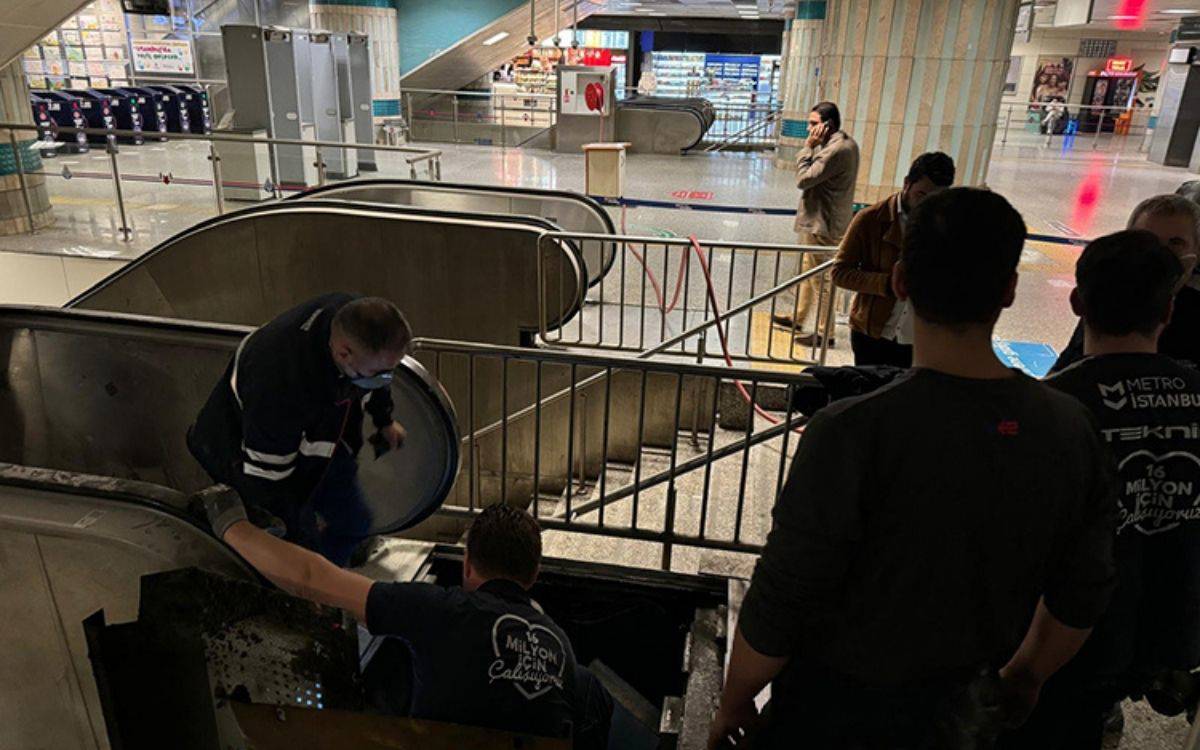 Bu fotoğrafta, İstanbul'daki bir metro istasyonunda yürüyen merdivenin onarım çalışmaları yapılırken görülüyor. Çalışma alanı güvenlik bariyerleriyle çevrilmiş durumda ve birkaç teknik personel yürüyen merdivenin içine eğilerek tamir işlemleri yapıyor. Çalışanlardan biri maskeli, diğer çalışanlar da merdivenin içinde ya da çevresinde bulunuyor. Arkada başka yolcular ve teknik ekip çalışanları yer alırken, bazı kişiler telefonla konuşuyor. İstasyonun iç kısmında çeşitli mağazalar ve otomatlar göze çarpıyor. Bu görüntü, metroda arıza sonrası yürütülen teknik müdahale sürecini yansıtıyor.