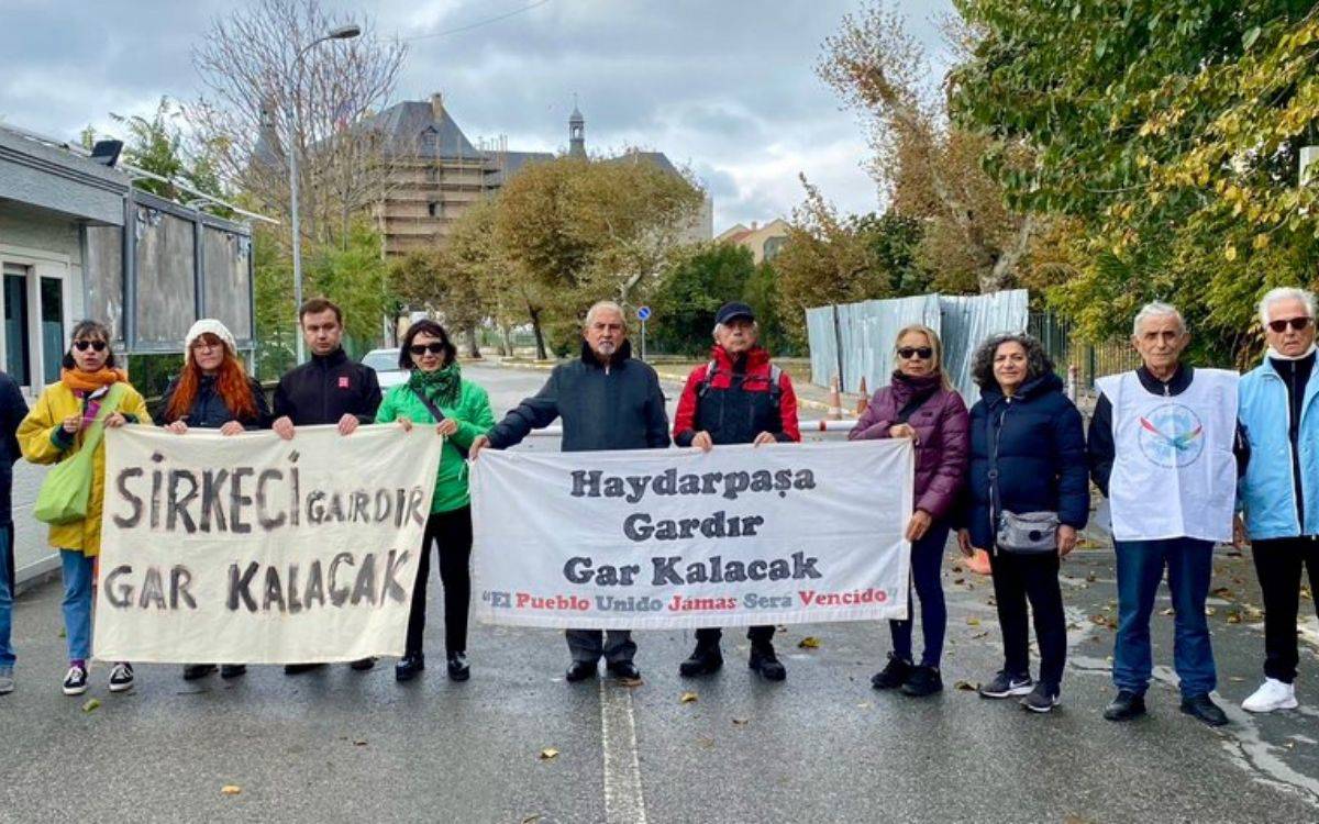 Bu fotoğrafta bir grup insan, ellerinde pankartlarla birlikte bir protesto veya destek etkinliğinde bulunuyor. Katılımcılar soğuk bir hava koşuluna uygun olarak giyinmiş. Arka planda ağaçlar, bazı binalar ve bir yapım çalışmasını düşündüren çevre düzenlemesi mevcut.