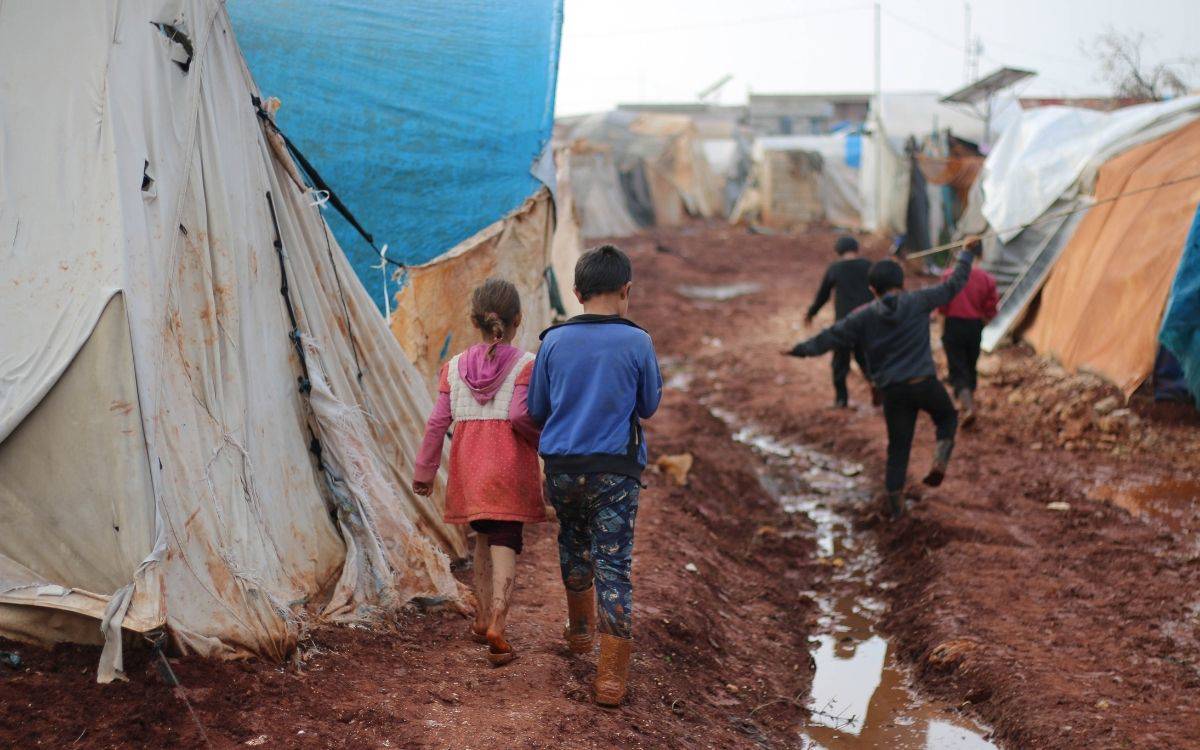 Bu fotoğrafta, çamurla kaplı bir alanda yürüyen çocuklar görülüyor. Çocukların ayaklarında çamurla kaplanmış botlar var ve üzerlerinde kışlık giysiler mevcut. Bulundukları alan, büyük ihtimalle bir mülteci kampı veya zorunlu barınma alanı; çünkü çevrede plastik örtülerle kaplanmış, eski ve yıpranmış çadırlar sıralanmış durumda. Çadırların kenarları kirlenmiş ve aşınmış. Yürüdükleri patika çamurlu, ıslak ve yer yer su birikintileri bulunuyor.