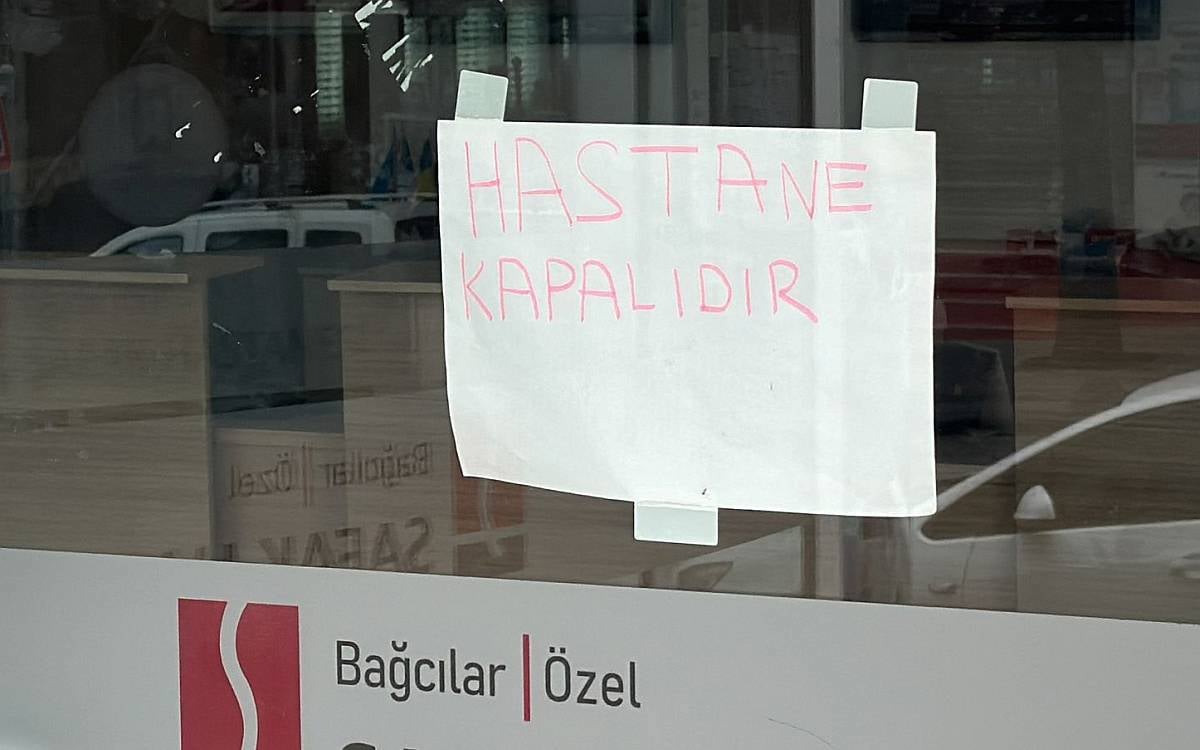 Fotoğrafta, bir hastanenin cam kapısına elle yazılmış bir duyuru asıldığı görülüyor. Beyaz bir kağıda kırmızı kalemle büyük harflerle yazılmış "HASTANE KAPALIDIR" ifadesi dikkat çekiyor. Yazı, bantlarla cama tutturulmuş. Camın arkasında hastanenin iç kısmı bulanık bir şekilde görülebiliyor; içeride masa veya mobilyaya benzeyen unsurlar bulunuyor. Camın alt kısmında ise "Bağcılar Özel" ifadesi ve bir logo seçilebiliyor. Fotoğraf, bir hastanenin hizmet dışı olduğunu açık bir şekilde duyurmak amacıyla hazırlanmış basit bir uyarı niteliği taşıyor.