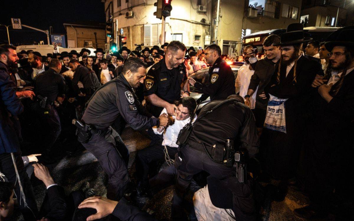 Bu görüntüde gece vakti bir sokakta toplanmış büyük bir grup insan görülüyor. Ön planda, polis memurları bir kişiyi yere doğru bastırarak kontrol altına almaya çalışıyor. Polislerin üzerinde resmi üniformaları ve ekipmanları var. Bu kişinin kıyafetleri, ortodoks Yahudi topluluğu üyelerini anımsatan geleneksel beyaz gömlek ve siyah pantolon gibi kıyafetlere benziyor.  Etrafta toplanmış kalabalık, olayları izliyor ve bazıları şaşkın, endişeli veya tepkili görünüyor. Birçoğu siyah takım elbise, beyaz gömlek ve geleneksel şapka gibi dini giysiler giymiş. Arkada bir bina ve birkaç sokak lambası görülebiliyor. Olay yeri kaotik bir atmosferi yansıtıyor ve polis ile siviller arasındaki bir gerginliği ifade ediyor.