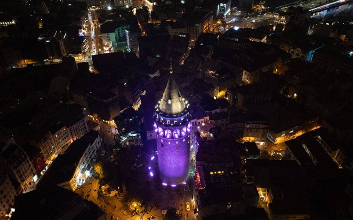 Bu görüntüde, gece vakti Galata Kulesi'nin yukarıdan çekilmiş bir fotoğrafı yer alıyor. Kule, mor tonlarında ışıklarla aydınlatılmış ve etrafındaki alanda da yoğun bir şekilde ışıklandırmalar göze çarpıyor. Galata Kulesi'nin etrafındaki dar sokaklar, çevresindeki tarihi yapılar ve meydan belirgin şekilde görülüyor. Etrafında yoğun bir insan hareketliliği olduğu anlaşılıyor. Daha uzakta, İstanbul'un diğer kısımları da sokak lambaları ve bina ışıkları ile karanlıkta parıldıyor. Arka planda köprüler ve suyun yansıması seçiliyor, bu da şehrin sahil şeridini işaret ediyor. Görüntü, tarihi ve modern İstanbul'un gece atmosferini etkileyici bir şekilde yansıtıyor.