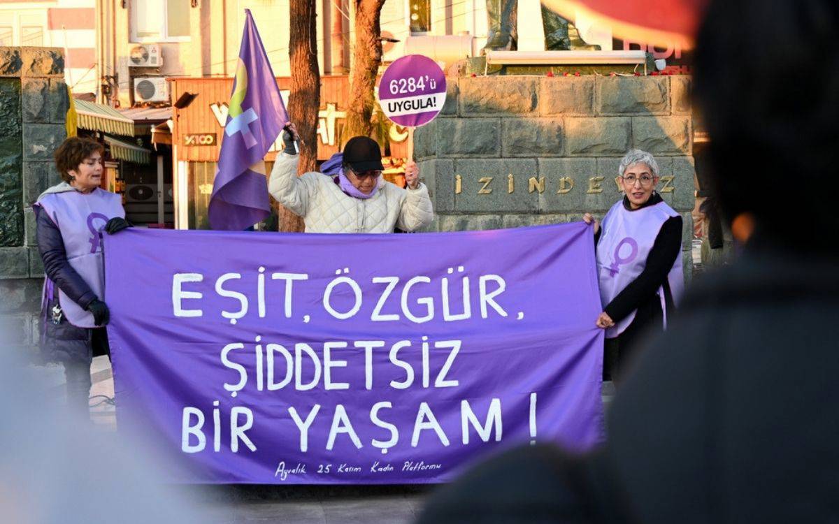 Fotoğrafta, üç kadın mor renkte kıyafetler ve aksesuarlar ile bir meydanda protesto gösterisi düzenliyor. Ellerinde büyük bir pankart var ve üzerinde büyük beyaz harflerle "Eşit, Özgür, Şiddetsiz Bir Yaşam!" yazıyor. Pankartın altında "Ayvalık 25 Kasım Kadın Platformu" ifadesi yer alıyor. Ortadaki kadın, bir elinde mor bir bayrak tutarken diğer elinde dairesel bir döviz taşıyor. Dövizde "6284’ü Uygula!" yazısı dikkat çekiyor. Arkada, taş bir duvar ve "İzindeyiz" yazılı bir anıt görülüyor. Çevrede ağaçlar ve bazı dükkanlar var. Protesto, kadın hakları ve şiddete karşı mücadeleye yönelik bir etkinliği temsil ediyor.