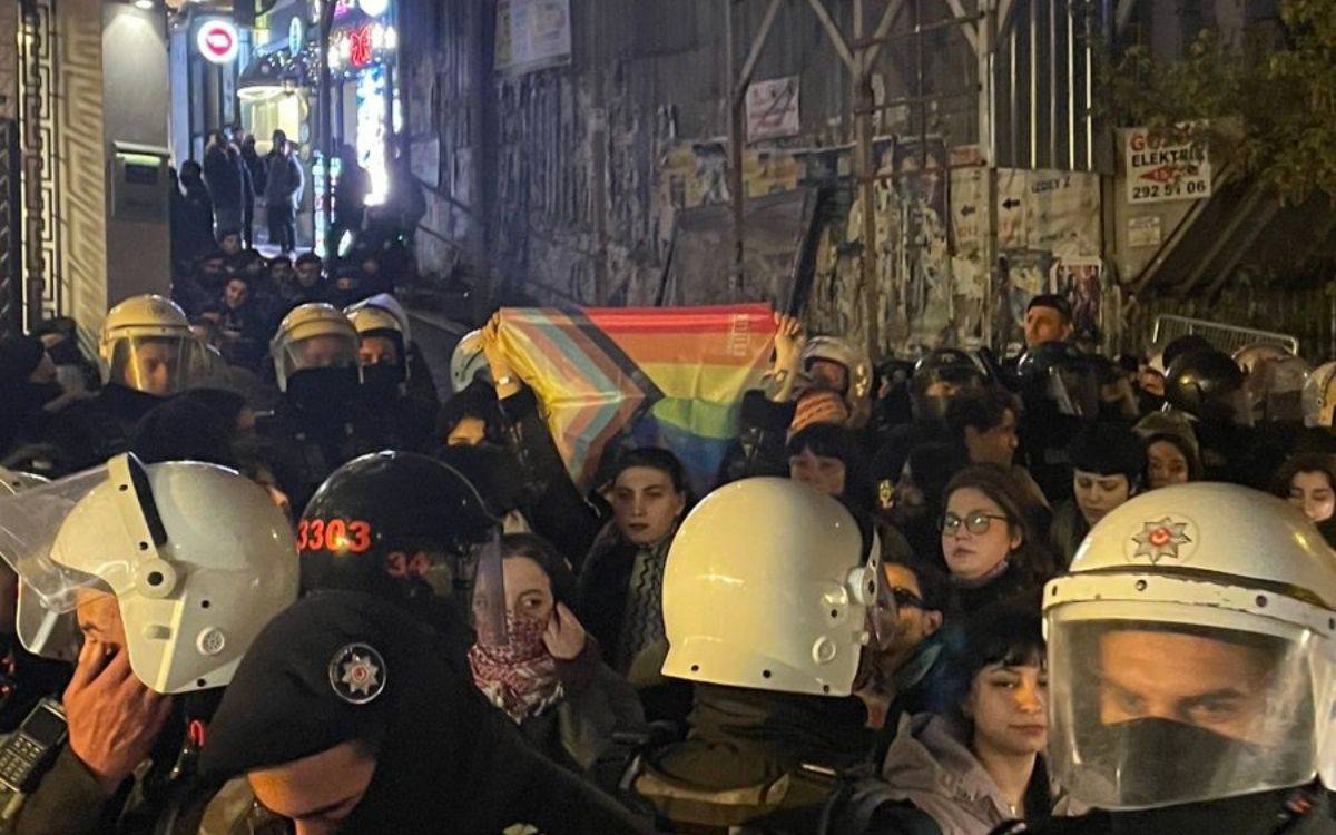 Fotoğrafta, gece saatlerinde dar bir sokakta yoğun bir polis ve protestocu kalabalığı görülüyor. Çevik kuvvet polisleri, kask ve koruyucu ekipmanlarla alanda güvenlik önlemi almış durumda. Polislerin çevrelediği alanda protestocular, ellerinde bir gökkuşağı bayrağı (LGBTQIA+ topluluğunu simgeleyen) tutarak eylemlerini sürdürüyor.