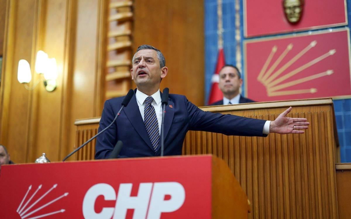 Fotoğrafta, Cumhuriyet Halk Partisi (CHP) Genel Başkanı Özgür Özel, partisinin grup toplantısında kürsüde konuşma yaparken görülüyor. Özel, siyah bir takım elbise ve çizgili bir kravat giyiyor. Elini yan tarafa doğru uzatarak vurgu yaparken, yüz ifadesi güçlü bir şekilde bir noktayı açıklamaya çalıştığını gösteriyor. Arkada, CHP logosunun ve Türk bayrağının bulunduğu mavi bir arka plan yer alıyor. Kürsünün önünde CHP'nin kırmızı zemin üzerine beyaz harflerle yazılmış logosu dikkat çekiyor. Fotoğraf, Özgür Özel'in kararlı ve etkileyici bir konuşma yaptığı anı yansıtıyor. Arkada bir kişi de izleyici pozisyonunda duruyor ve toplantının ciddiyetini pekiştiriyor.