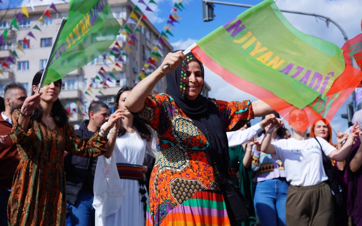 Fotoğraf bir mitingden. Önde bir kadın elinde Jin, Jiyan, Azadi yazan bir bayrak ile dans ediyor. Arkasında kalabalık bir kadın grubu var.