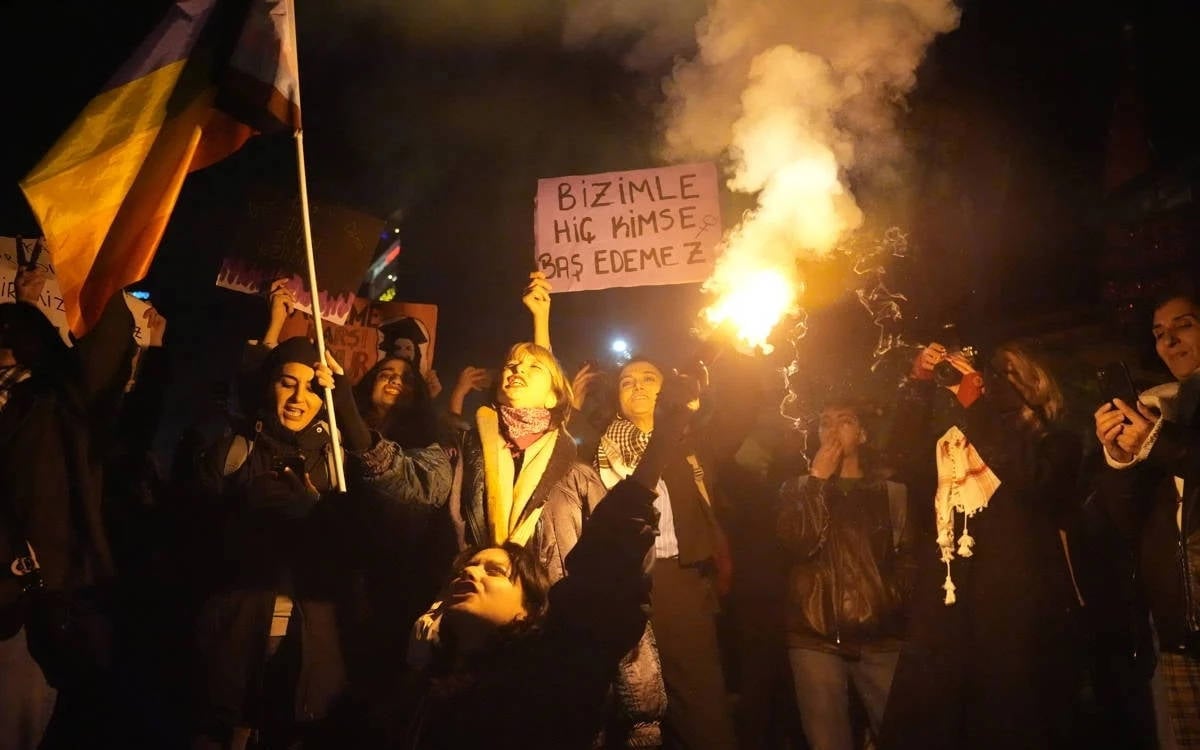 Bu fotoğrafta, bir grup insan gece saatlerinde bir protesto ya da yürüyüş sırasında görülüyor. Kalabalığın içinde kadınlar çoğunlukta ve bazı kişiler ellerinde pankartlar tutuyor. Öne çıkan pankartlardan birinde "Bizimle Hiç Kimse Baş Edemez" yazısı dikkat çekiyor. Gruptan biri meşale taşıyarak dikkat çeken bir ışık kaynağı oluşturuyor. Sol tarafta gökkuşağı renklerinde bir bayrak dalgalanıyor. Katılımcılar arasında coşkulu ve kararlı bir atmosfer hâkim. Arkadaki karanlık ortam, meşale ışığı ve pankartlar sahneyi aydınlatıyor. Bu, bir dayanışma veya hak savunusuna yönelik bir etkinlik gibi görünüyor.