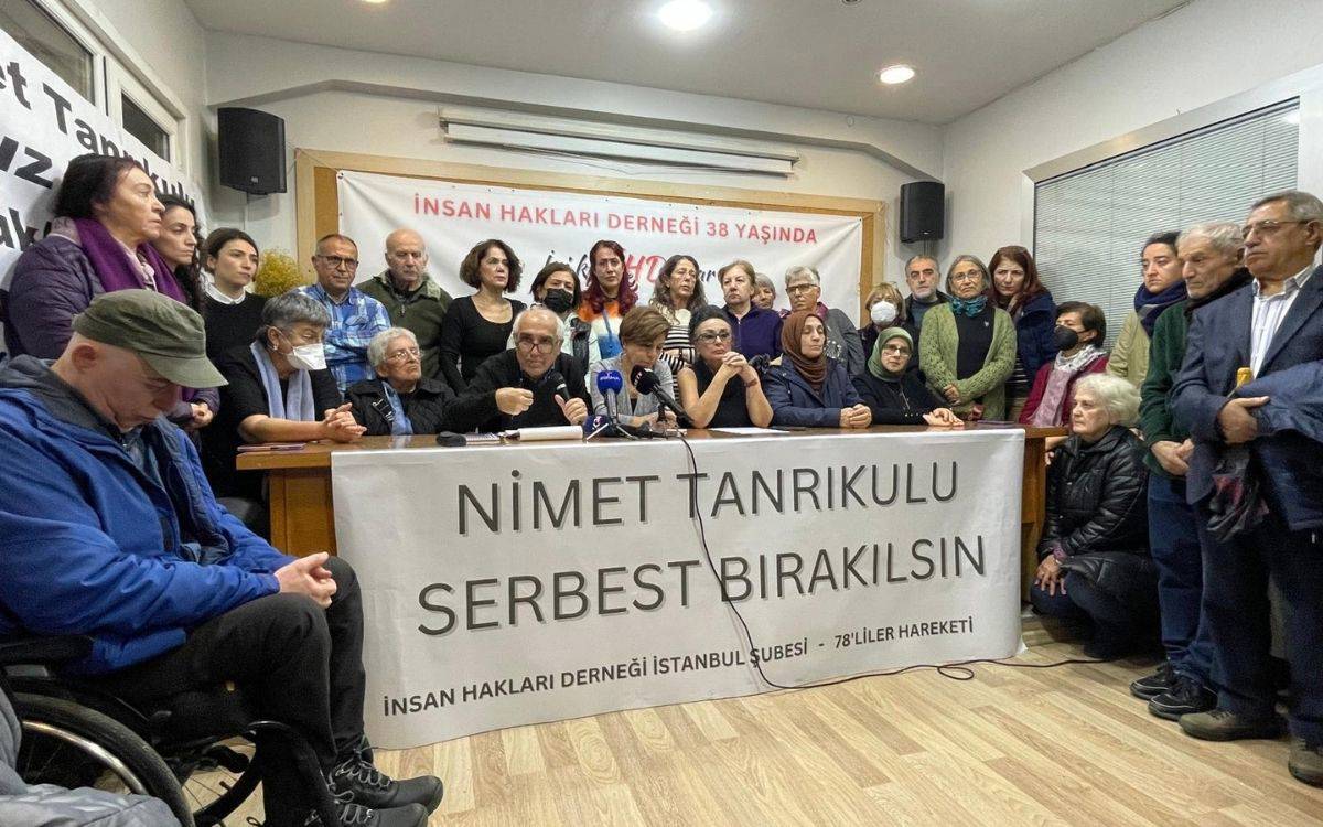 Bu fotoğrafta bir grup insan, "Nimet Tanrıkulu Serbest Bırakılsın" yazılı bir pankartın arkasında toplanmış şekilde görülüyor. Fotoğraf, bir basın açıklaması veya toplantısı sırasında çekilmiş gibi görünüyor. Arka planda "İnsan Hakları Derneği 38 Yaşında" yazılı bir afiş yer alıyor. Masada mikrofonlar bulunuyor, bu da toplantının medyaya açık bir etkinlik olduğunu gösteriyor. Katılımcılar arasında farklı yaşlardan kadın ve erkekler yer alıyor. Bazıları oturmuş, bazıları ayakta duruyor. Genel atmosfer ciddi ve dayanışma odaklı.