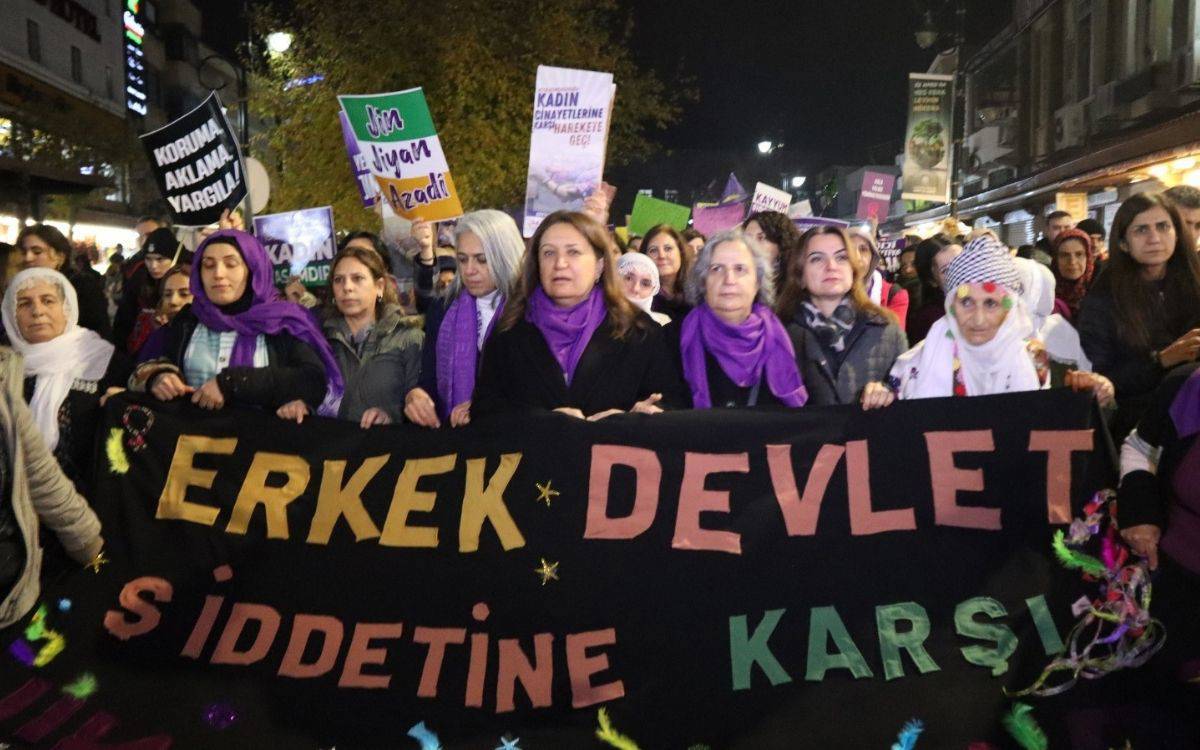 Bu fotoğrafta, gece saatlerinde bir protesto yürüyüşü görülüyor. Kadınlar, ellerinde pankartlar ve büyük bir siyah pankart taşıyor. Pankart üzerinde büyük harflerle "ERKEK DEVLET ŞİDDETİNE KARŞI" yazıyor. Pankartı taşıyan kadınların birçoğunun boynunda mor atkılar ve şallar bulunuyor; bu da etkinliğin kadın hakları veya kadın dayanışmasına yönelik bir protesto olduğunu gösteriyor.  Arkada diğer protestocular çeşitli pankartlar taşıyor. Öne çıkan pankartlardan bazıları şunları içeriyor: "Jin, Jiyan, Azadî" ve "Kadın Cinayetlerine Tahammülümüz Yok" gibi mesajlar. Kalabalık bir grup, kararlı bir şekilde yürüyüşe devam ediyor. Eylem alanı yoğun bir şekilde aydınlatılmış, arka planda ağaçlar ve sokak lambaları dikkat çekiyor.  Katılımcılar arasında her yaştan kadın bulunuyor, bu da etkinliğin kapsayıcı bir protesto olduğunun altını çiziyor. Genel atmosfer dayanışma, kararlılık ve toplumsal farkındalık yaratma isteğiyle dolu.