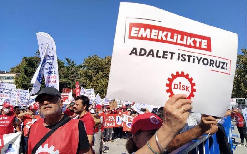 Bu fotoğrafta, bir grup insan emeklilik haklarıyla ilgili bir protesto gerçekleştirmektedir. Kalabalık, açık bir alanda toplanmış ve çeşitli pankartlar taşımaktadır. Ön planda, bir kişinin tuttuğu pankartta büyük harflerle "EMEKLİLİKTE ADALET İSTİYORUZ!" yazısı dikkat çekmektedir. Pankartın altında DİSK (Devrimci İşçi Sendikaları Konfederasyonu) logosu yer almaktadır. Katılımcıların çoğu kırmızı renkli kıyafetler giymiş ya da kırmızı şapkalar takmıştır. Arka planda ise farklı sendikaların isimlerinin yazılı olduğu bayrak ve pankartlar görülmektedir. Protesto, organize bir şekilde ve hak arayışı teması etrafında gerçekleştirilmiş gibi görünmektedir. Etkinlikte sıcak bir hava olduğu izlenimi, katılımcıların güneş altında bulunmasından anlaşılmaktadır.