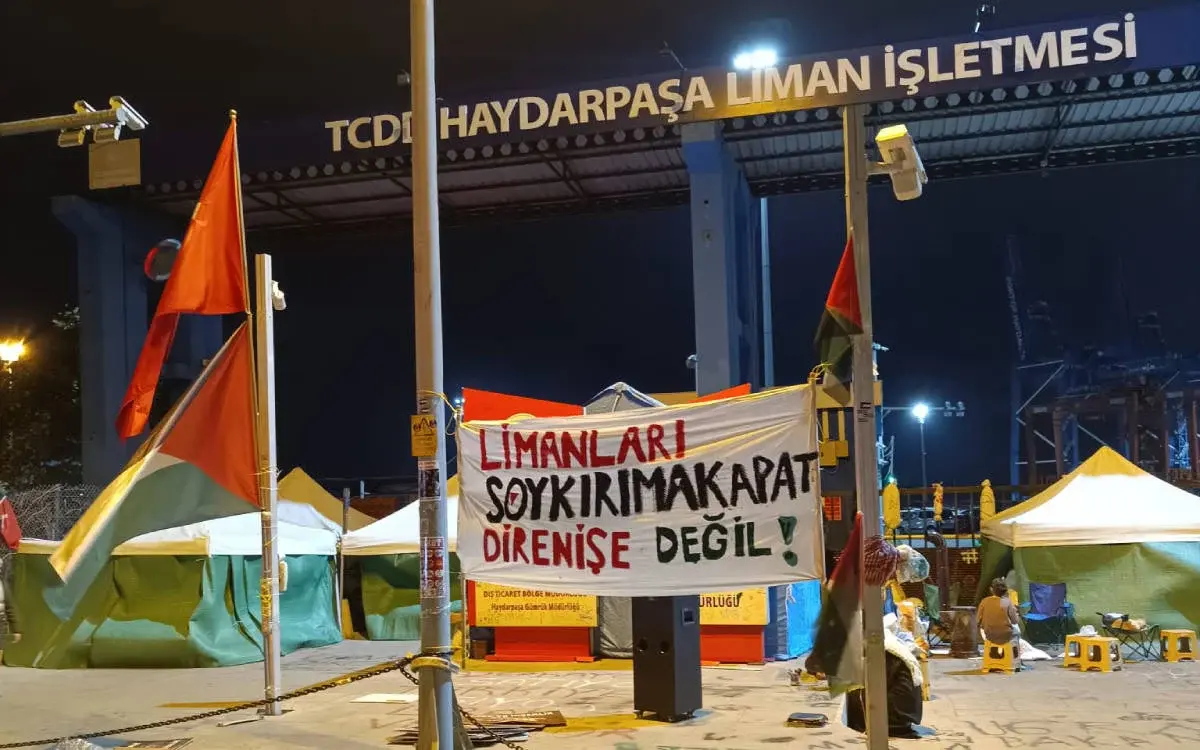 Bu fotoğraf, gece saatlerinde bir liman alanında çekilmiş. Arka planda, "TCDD Haydarpaşa Liman İşletmesi" yazısı yer alan bir giriş kapısı görülüyor. Girişin önüne pankartlar ve çadırlar kurulmuş. Beyaz bir pankartta büyük harflerle "Limanları Soykırıma Kapat, Direnişe Değil!" ifadesi yer alıyor. Pankart, protestonun ana mesajını yansıtıyor. Ayrıca, çevrede kırmızı bayraklar ve Filistin bayrakları dikkat çekiyor.