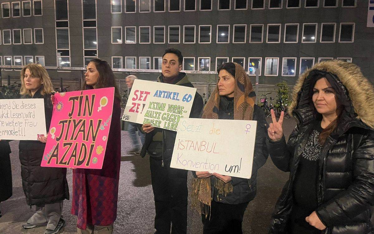 Bu fotoğraf, bir protesto etkinliğinde yer alan bir grup kadını ve bir erkeği göstermektedir. Katılımcılar, çeşitli pankartlar tutarak dikkat çekiyorlar. Pankartlarda hem Almanca hem de Kürtçe ifadeler bulunuyor. Örneğin, ortadaki kişi "Jin, Jiyan, Azadî" yazılı parlak pembe bir pankart tutarken, diğer katılımcılar İstanbul Sözleşmesi'nin uygulanması ve şiddete karşı talepler içeren Almanca mesajlar taşıyor.  Katılımcılar arasında farklı yaş gruplarından bireyler yer alıyor ve bir kısmı kışlık giysiler giymiş. Arka planda, çok katlı bir binanın camlı cephesi ve az sayıda insan hareketi görülüyor.