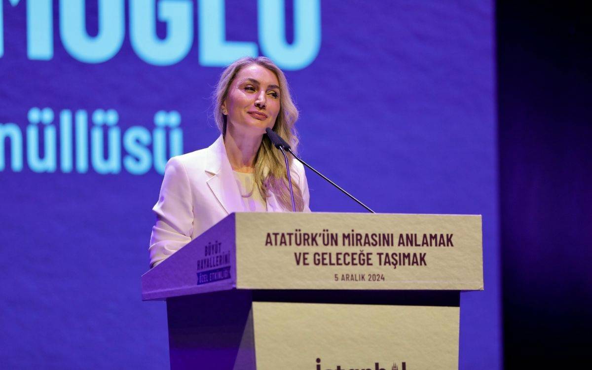 Fotoğrafta, Dr. Dilek Kaya İmamoğlu, sahnede bir konuşma yaparken görülüyor. Mor bir arka planın önünde yer alan kürsüde, etkinliğin adı olan "Atatürk'ün Mirasını Anlamak ve Geleceğe Taşımak" yazısı dikkat çekiyor. Kürsünün üzerinde aynı zamanda etkinliğin tarihi olan "5 Aralık 2024" ifadesi yer alıyor. Dr. İmamoğlu, beyaz bir takım elbise giymiş, zarif ve resmi bir görünüm sergiliyor. Yüzünde sakin ve kararlı bir ifade var, konuşmasını yaparken topluluğa hitap ediyor. Arka planda ise büyük puntolarla yazılmış metinlerden “Gönüllüsü” kelimesi seçilebiliyor. Bu fotoğraf, toplumsal cinsiyet eşitliği ve kadın haklarına dair önemli bir etkinlikte yapılan konuşmayı temsil ediyor.