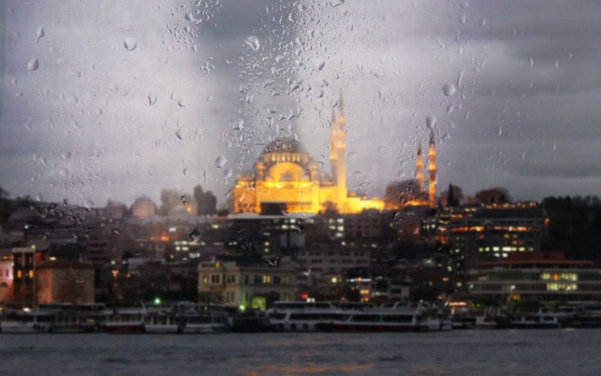 Fotoğrafta, yağmurlu bir havada İstanbul’un ikonik bir görüntüsü yer alıyor. Cam üzerinde yağmur damlaları odakta ve bu damlaların arkasında, aydınlatılmış bir cami silueti belirgin şekilde görülüyor. Arka planda İstanbul’un tarihi dokusunu yansıtan yapılar, deniz ve kıyıda dizili vapurlar dikkat çekiyor. Gökyüzü gri bulutlarla kaplı, bu da yağışlı ve melankolik bir atmosfer yaratıyor. Fotoğraf, İstanbul’un yağmurlu bir günündeki huzurlu ve mistik havasını yansıtıyor.