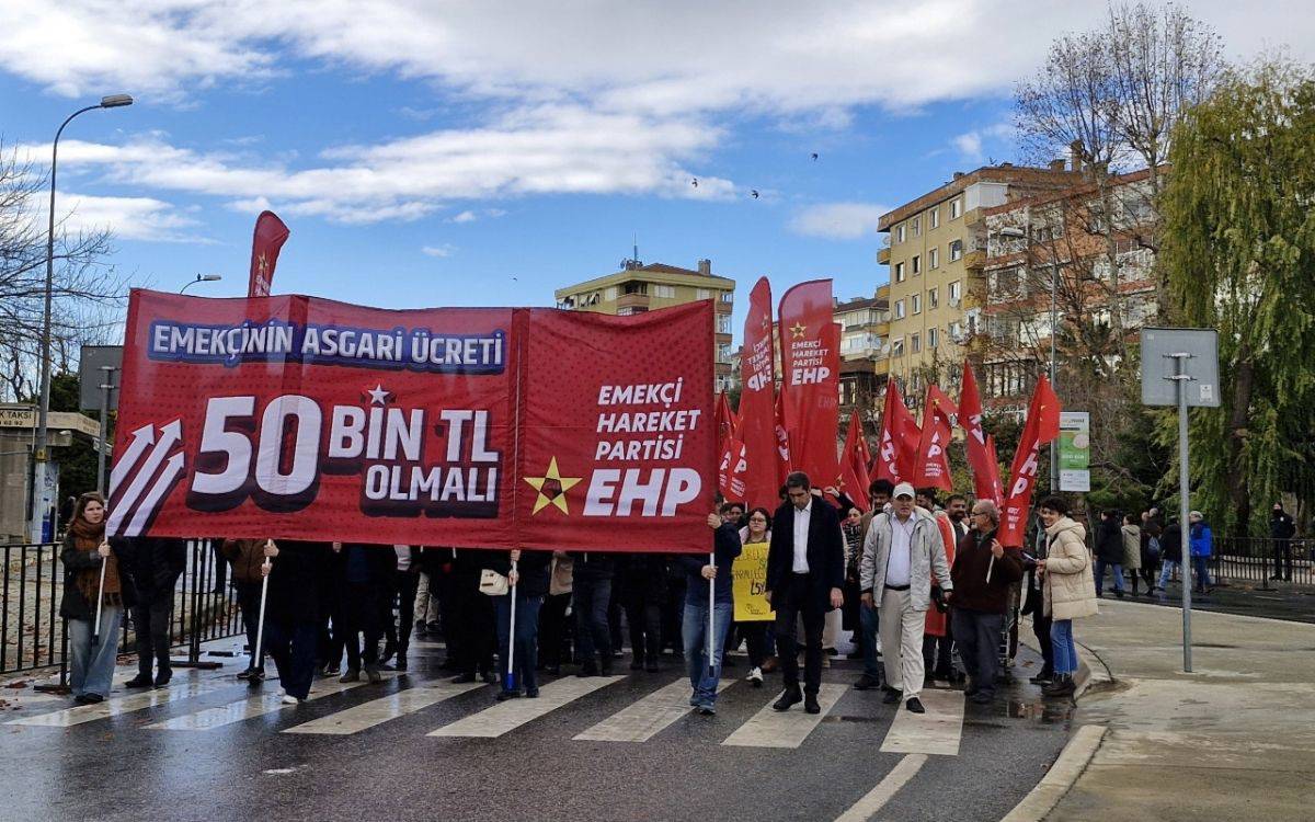 Fotoğrafta, bir grup insan bir yürüyüş sırasında ellerinde pankart ve bayraklarla görülüyor. Kalabalık, büyük bir kırmızı pankart taşımakta; üzerinde "Emekçinin Asgari Ücreti 50 Bin TL Olmalı" yazısı dikkat çekiyor. Pankartın solunda beyaz ok işaretleri ve sağında "Emekçi Hareket Partisi (EHP)" logosu ile yazısı yer alıyor. Arkada kırmızı renkli birçok bayrak taşınıyor, bayrakların üzerinde yine "EHP" yazıları ve yıldız sembolleri var.  Katılımcılar, bir yaya geçidinde yürüyerek taleplerini dile getiriyor. Hava açık ama serin görünüyor, bazı kişiler mont giymiş. Çevrede ağaçlar, apartmanlar ve açık alanlar fotoğrafa dahil. Bu görüntü, bir siyasi hareketin ekonomik taleplerini dile getiren organize bir protesto veya yürüyüşü temsil ediyor.