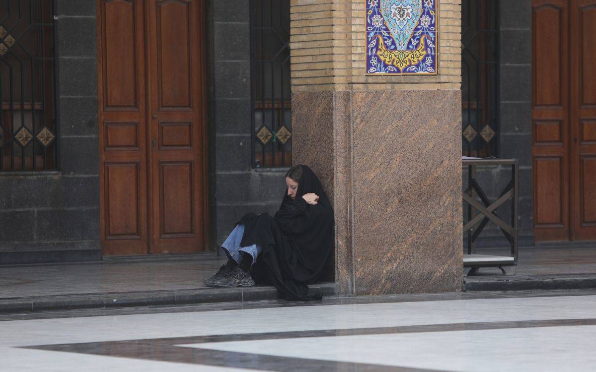 Bu fotoğraf, bir mimari yapının dışındaki bir kadını tasvir ediyor. Kadın, açık bir alanda, bir sütunun köşesine yaslanmış şekilde oturuyor. Siyah bir çarşaf giymiş ve başını örten bir şekilde, kollarını kendine sararak içe dönük bir duruş sergiliyor. Bu haliyle sakin veya hüzünlü bir izlenim bırakıyor.  Arka planda, tuğla ve koyu taşlarla kaplanmış bir duvar bulunuyor. Duvar üzerinde geleneksel motiflerle süslenmiş renkli bir çini dikkat çekiyor. Yan tarafta ise ahşap kapılar ve demir parmaklıklarla çevrili pencereler yer alıyor. Zeminde parlak taş kaplama görülüyor ve yerin düzenli geometrik desenlerle döşendiği fark ediliyor.  Fotoğraf, yalnızlık ve düşünceli bir ruh hali hissi uyandırırken, aynı zamanda mimari detaylar sayesinde mekânın kültürel veya dini bir bağlam taşıdığına işaret ediyor.