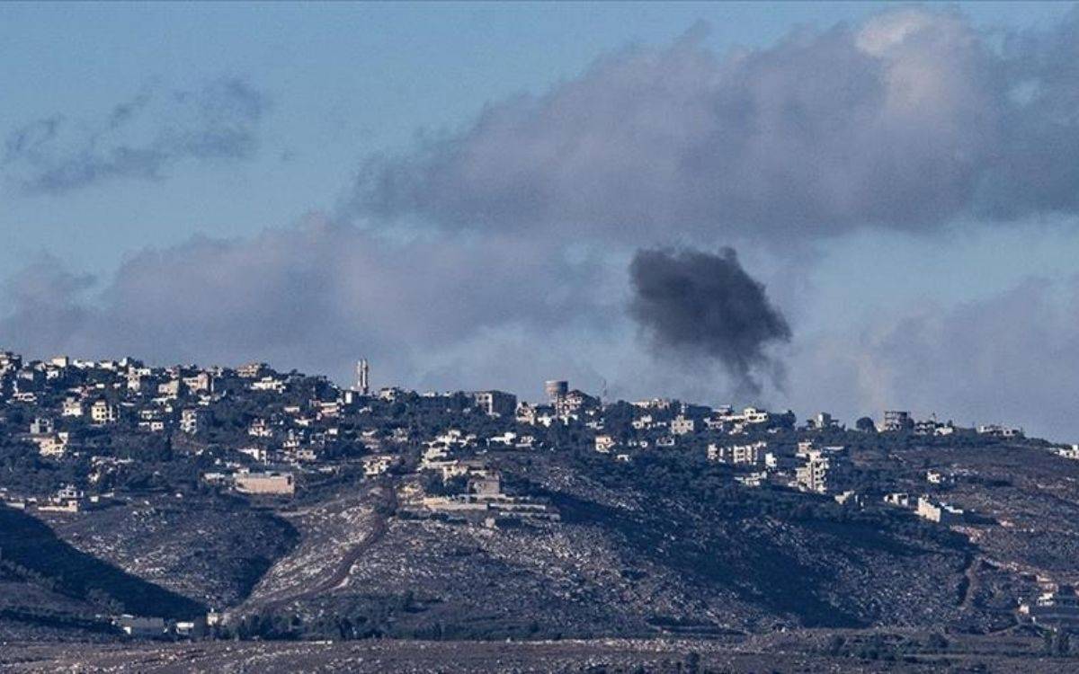 Bu fotoğrafta bir tepe üzerinde yer alan bir yerleşim alanı görülüyor. Tepenin üzerinde çok sayıda bina dikkat çekiyor ve bu binalar genellikle yerleşim amaçlı kullanılan küçük yapılara benziyor. Fotoğrafın sağ tarafında, gökyüzüne doğru yükselen yoğun bir siyah duman bulutu dikkat çekiyor. Bu duman, bir patlama ya da yangın sonucunda oluşmuş gibi görünüyor. Arka planda ise bulutlu bir gökyüzü var, ancak hava açık ve manzara net şekilde görülebiliyor.