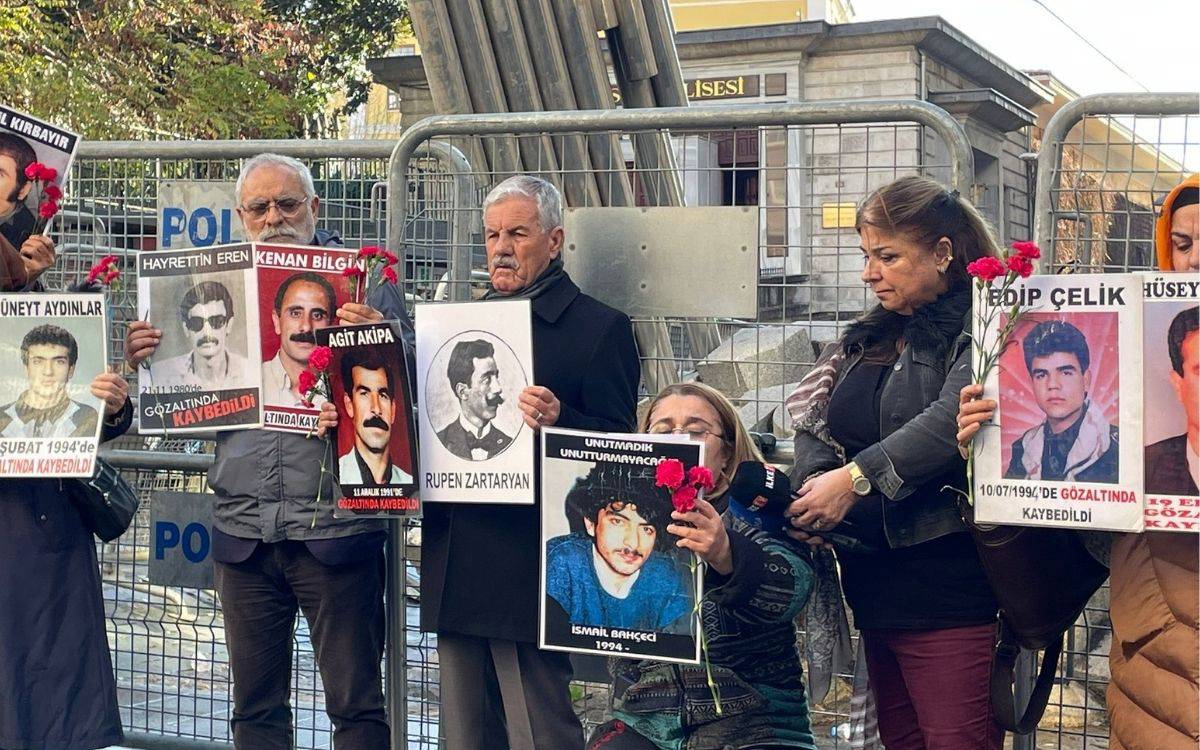 Bu fotoğrafta bir grup insan, ellerinde kayıp yakınlarının fotoğraflarını ve karanfiller tutarak bir protesto veya anma etkinliği düzenliyor. Arkalarında polis barikatları görülüyor. Her bir kişi, farklı bir kaybı temsil eden portre ve yazılı pankartlar taşıyor. Fotoğrafların altında "Gözaltında kaybedildi" gibi ifadeler yer almakta. Katılımcılar genellikle ciddi ve hüzünlü bir ifadeye sahip. Bu görüntü, kayıpların unutulmaması ve adalet talebi için yapılan bir etkinliği yansıtıyor gibi görünüyor.