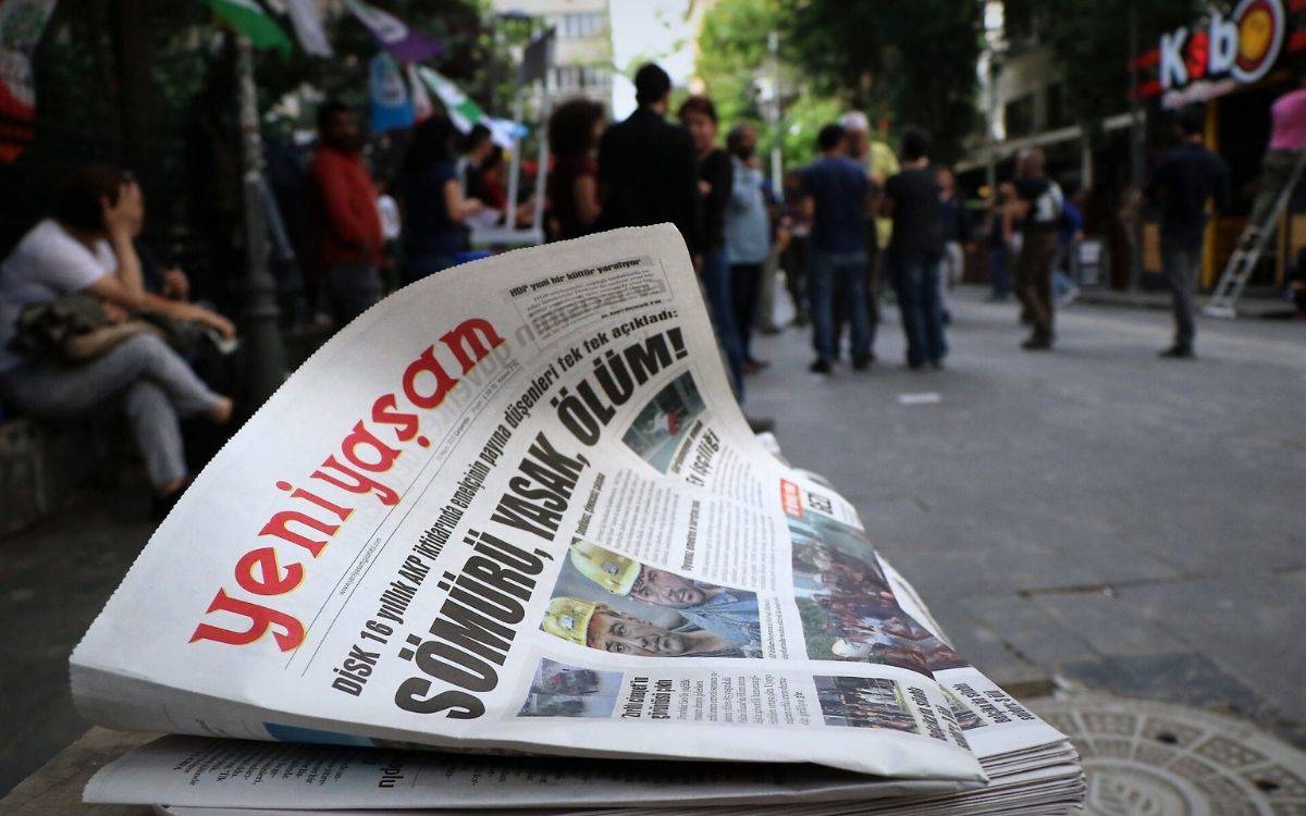 Fotoğrafta Yeni Yaşam gazetesinin bir sayısı bulunuyor. Arkada sokakta yürüyen insanlar var.