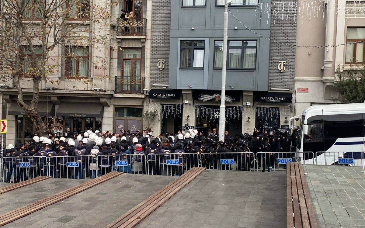 Bu fotoğraf, bir şehir meydanında çekilmiş ve yoğun bir polis varlığını göstermektedir. Görselde, polis bariyerleriyle çevrilmiş bir alanın önünde çok sayıda polis memuru görülüyor. Polislerin çoğunun kask taktığı ve bazı memurların tam teçhizatlı olduğu fark ediliyor. Arka planda, bir otel ve çevresinde çeşitli işletmeler yer alıyor. Otelin tabelasında "Galata Times" yazısı okunabiliyor. Binanın üst katlarındaki balkonlardan birkaç kişi meydandaki durumu izliyor gibi görünüyor. Meydanın ön kısmında ahşap banklar, arka kısımda ise park edilmiş bir polis otobüsü dikkat çekiyor. Meydanda bir olayın güvenlik önlemleri kapsamında kontrol altına alınmaya çalışıldığı izlenimi veriyor.