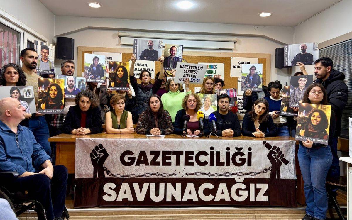 Fotoğrafta, bir grup insan bir masa etrafında toplanmış durumda ve çeşitli pankartlar ile afişler tutuyor. Masanın önünde "Gazeteciliği" yazılı bir pankart yer almakta, yanında ise bir yumruk sembolü dikkat çekiyor. İnsanların ellerinde tutulan afişlerde, tutuklu veya hedef alınmış gazetecilerin fotoğrafları yer almakta. Arkada, duvara asılmış farklı yazılar dikkat çekiyor; bunlar arasında "Gazetecileri Serbest Bırakın", "İşkence İnsanlık Suçudur" ve "Herkes Farklı Herkes Eşit" gibi mesajlar bulunuyor.  Masada oturanlar, ellerinde metin tutan ve konuşan kişilerle birlikte bir basın açıklaması yapıyor gibi görünüyor. Çevrede toplanan diğer insanlar bu açıklamaya destek verir nitelikte bir duruş sergiliyor. Bu kare, basın özgürlüğü ve gazetecilere yönelik saldırılara dikkat çekmeyi amaçlayan bir dayanışma etkinliğini temsil ediyor.