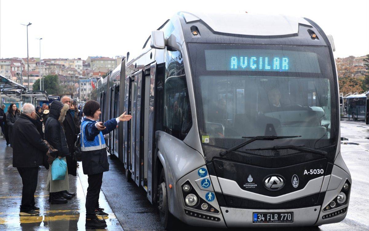 Fotoğrafta, bir metrobüs durağında bekleyen yolcular ve görevini yapan bir denetim amiri görülmektedir. Önde, mavi yelek giymiş bir kadın görevli (denetim amiri), metrobüs yolcularını yönlendirmek için elini uzatmış bir hareket yapmaktadır. Metrobüs üzerinde "Avcılar" yazısı bulunmakta, aracın ön kısmında İETT ve İstanbul Büyükşehir Belediyesi logoları dikkat çekmektedir.  Arka planda, durağa doğru sıraya girmiş çeşitli yaşlardan yolcular görülmekte. Hafif yağmurlu bir hava olduğu, zemindeki yansımalar ve insanların giydiği montlardan anlaşılmaktadır. Fotoğraf, hem toplu taşımada işleyen düzeni hem de kadınların bu sektördeki aktif rolünü yansıtıyor.