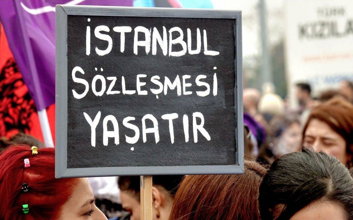 Fotoğrafta, bir protesto veya yürüyüş sırasında çekilmiş bir kare görülmektedir. Görselde, kalabalığın içinde bir kişi elinde siyah bir pankart tutmaktadır. Pankartın üzerinde beyaz harflerle "İstanbul Sözleşmesi Yaşatır" yazmaktadır. Pankart, kadına yönelik şiddetle mücadele ve İstanbul Sözleşmesi’nin önemine dikkat çeken bir mesaj içermektedir. Arka planda çeşitli kişilerin silüetleri görülmekte, pankart ve mesaj ön planda vurgulanmaktadır. Katılımcılar arasında mor renkli bayraklar da dikkat çekmektedir ve genel atmosfer, hak talebinde bulunan bir dayanışma havasını yansıtmaktadır.