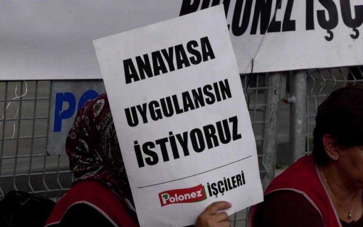 Fotoğrafta bir kadın, üzerinde "Anayasa uygulansın istiyoruz" yazan bir pankart tutuyor. Pankartın alt kısmında "Polonez İşçileri" ifadesi yer alıyor. Kadın, pankartın arkasında yüzünü gizlemiş, ancak başörtüsü dikkat çekiyor. Fotoğrafın arka planında, "Polonez" yazısı ve bir çit detayı görülüyor. Kadının yanındaki bir diğer kişi ise kırmızı önlük giyiyor, bu da fabrika işçilerini temsil eden bir kıyafet olabilir. Görüntü, işçilerin anayasal haklarını talep eden bir protesto eylemini yansıtıyor.