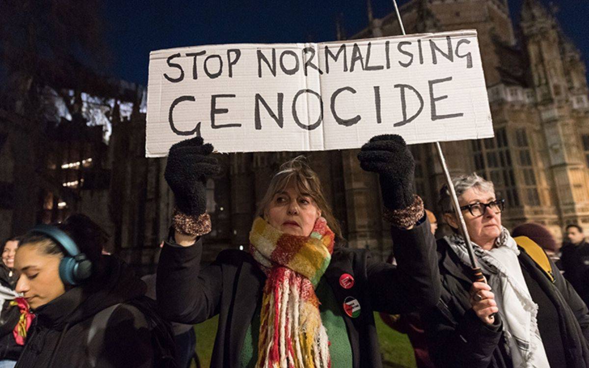 Fotoğrafta bir grup insan bir protesto sırasında görülmektedir. Ön plandaki bir kadın, üzerinde "Stop Normalising Genocide" (Soykırımı Normalleştirmeyi Durdurun) yazan bir pankart tutmaktadır. Kadın, boynunda renkli bir atkı ve siyah eldiven giymiştir. Arkasında başka kişiler de bulunmaktadır; biri gözlük takıyor ve diğerleri dikkatle etrafı izliyor. Fotoğraf, muhtemelen gece vakti çekilmiş olup, arka planda bir bina veya tarihi bir yapı görünmektedir. Genel atmosfer, bir toplumsal protesto ya da farkındalık etkinliğini çağrıştırmaktadır.