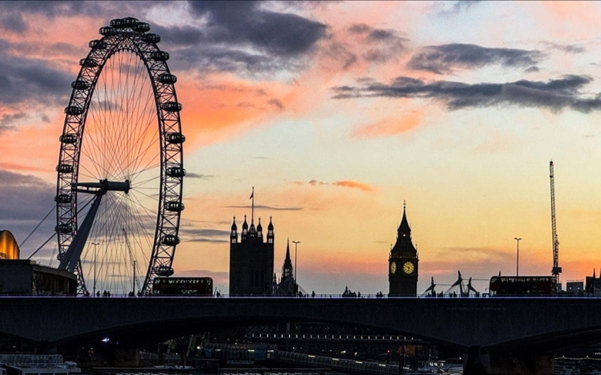 Bu görsel, İngiltere'nin başkenti Londra'dan bir manzara sunuyor. Arka planda gün batımında pastel tonlarında bir gökyüzü yer alıyor. Görüntünün solunda, Londra'nın ikonik dönme dolabı olan "London Eye" belirgin bir şekilde görülüyor. Sağda ise Big Ben saat kulesi ve Parlamento Binası’nın silueti bulunuyor. Önde bir köprü, üstünde otobüsler ve yayalarla birlikte gözüküyor. Köprünün alt kısmında Thames Nehri'nin kıyıları hafifçe seçiliyor. Genel olarak, bu sahne Londra'nın meşhur sembollerini ve romantik bir gün batımı atmosferini yansıtıyor.