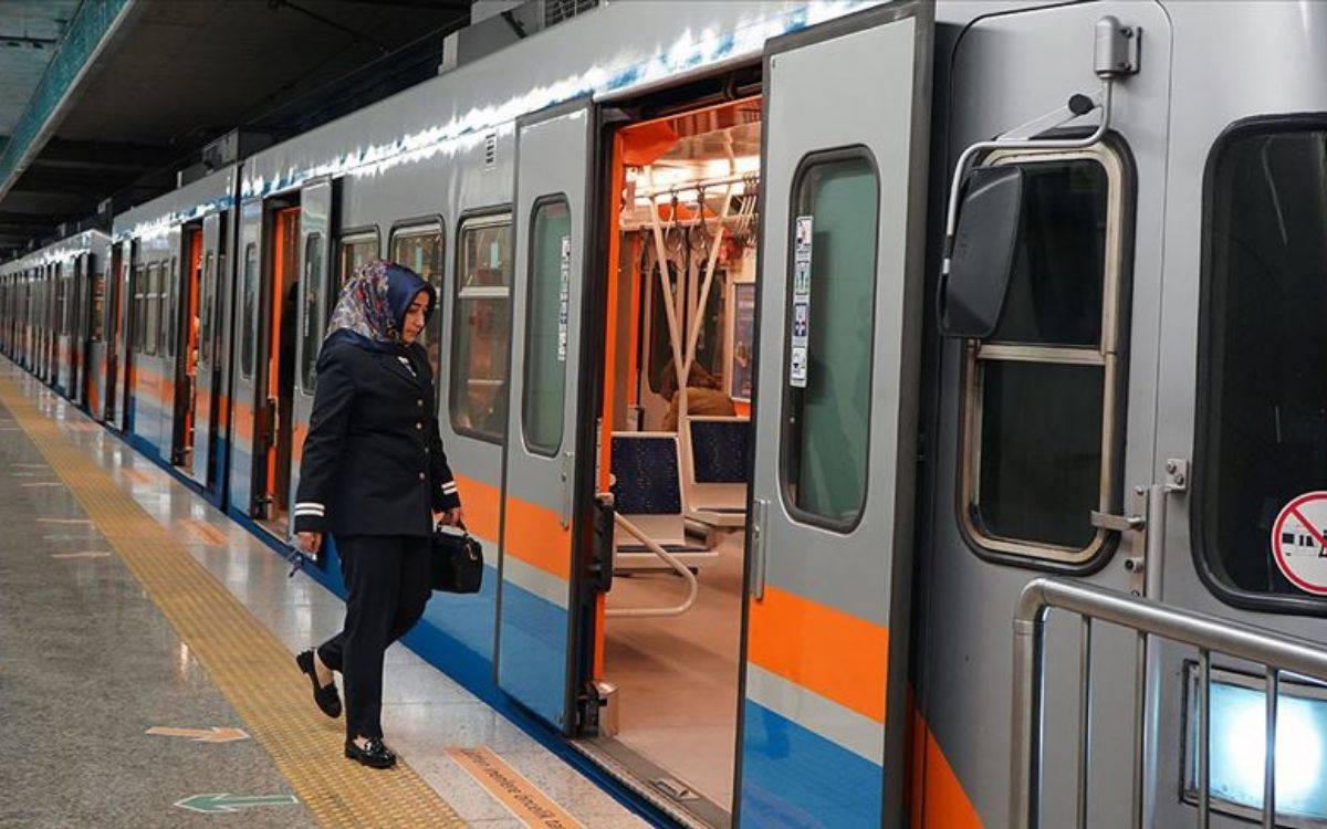 Fotoğrafta, bir kadın metro istasyonunda bir trenin kapısının önünde duruyor. Kadın, koyu lacivert bir üniforma giymiş ve başörtüsü takıyor. Elinde bir çanta taşıyor ve dikkatlice trenin kapısına doğru bakıyor. Metro aracı turuncu ve mavi şeritlere sahip gri renkte, kapıları açık durumda. Platformda sarı renkli bir güvenlik şeridi mevcut, bu da yolcuların güvenli mesafeyi korumasını sağlamak için kullanılıyor. Arka planda metro aracının iç kısmı görülüyor ve boş koltuklar dikkat çekiyor. Fotoğraf, bir metro istasyonunda günlük yaşamın bir anını yansıtıyor.