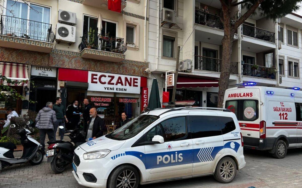 Bu görsel, Türkiye'de bir apartmanın önündeki hareketli bir anı göstermektedir. Görselde ön planda bir polis aracı ve bir ambulans dikkat çekiyor. Her iki araç da olay yerinde görevde gibi görünüyor. Eczane tabelası bulunan bir dükkânın önünde birkaç kişi bir araya toplanmış; bazıları birbirleriyle konuşuyor, bazılarıysa olayı izliyor gibi görünüyor.