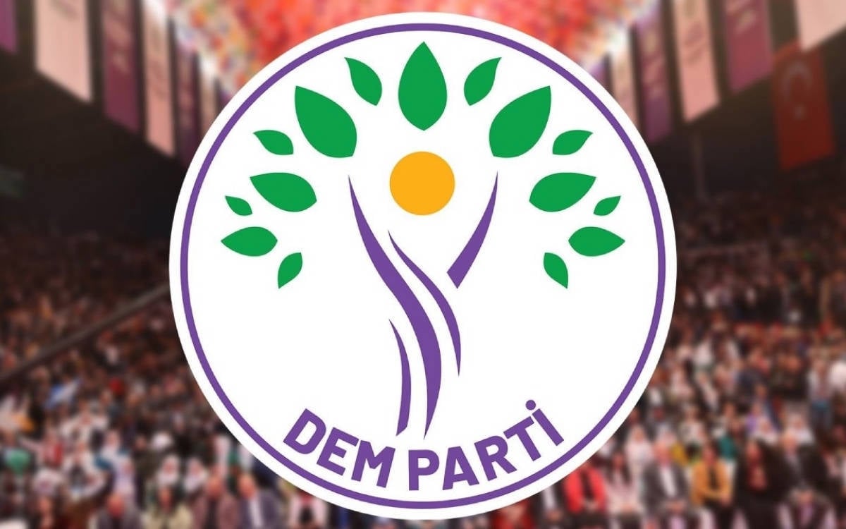 u fotoğrafta, Halkların Eşitlik ve Demokrasi Partisi'ne (DEM Parti) ait olduğu anlaşılan bir logo yer alıyor. Logo, bir ağacı andıran semboller içeriyor; mor ve yeşil tonlarıyla tasarlanmış. Yeşil yapraklar, doğayı veya canlılığı temsil ederken, merkezde bir daire (muhtemelen bir güneşi simgeliyor) yer almakta. Logo, mor renkle yazılmış "DEM PARTİ" ifadesiyle tamamlanıyor. Arka planda, bulanık bir şekilde kalabalık bir toplantı ya da kongre ortamı görünüyor.