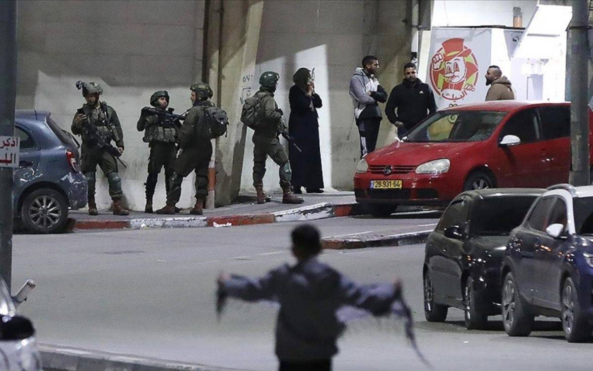 Bu fotoğrafta, gece saatlerinde bir şehir sokağı görülüyor. Ön planda bulanık bir figür yer alıyor ve bu figür kollarını iki yana açmış durumda. Arka planda ise silahlı askerler bir grup insanın önünde duruyor. Askerler tam teçhizatlı ve çevrede devriye yapıyor gibi görünüyor. Sokakta kırmızı bir araba ve birkaç park etmiş başka araç da dikkat çekiyor. Fotoğraf genel olarak bir çatışma veya kontrol noktası sahnesini andırıyor. Ayrıca, bir fast-food restoranının tabelası da arka planda seçilebiliyor. Atmosfer gerilim dolu ve askerlerin varlığı bölgedeki güvenlik durumuna dair bir ipucu veriyor.