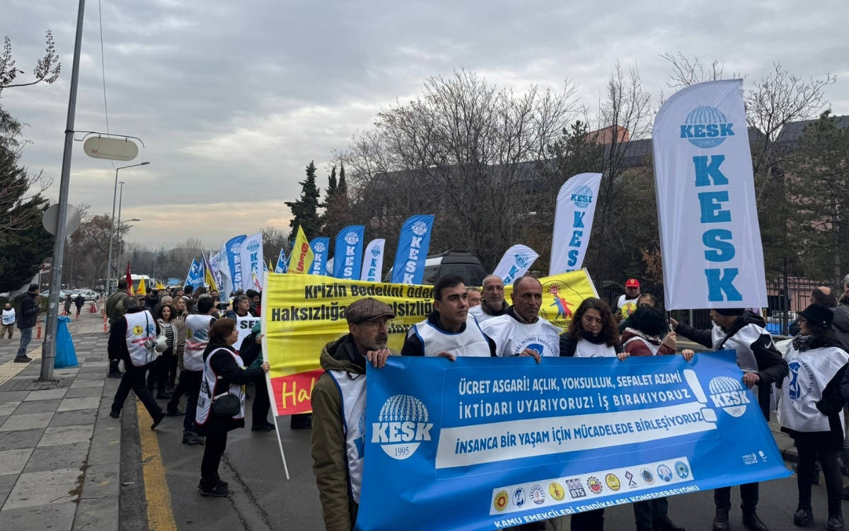 Fotoğraf kamu emekçilerinin Ankara'daki yürüyüşünden. Kalabalık grup önlerinde “Ücret Asgari! Açlık, Yoksulluk, Sefalet Azami İktidarı Uyarıyoruz! İş bırakıyoruz insanca bir yaşam için mücadelede birleşiyoruz” yazılı pankart ile yürüyor.