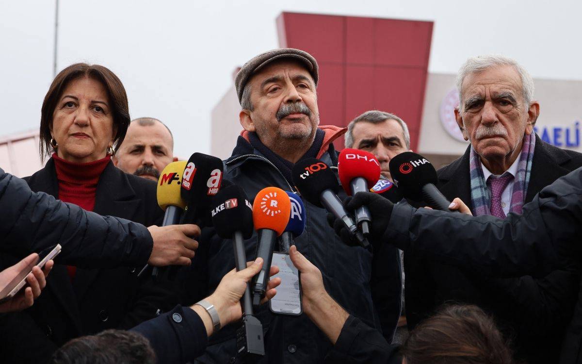 Bu görselde, resmi bir ortamda iki kişinin el sıkıştığı bir an görülmektedir. Soldaki kişi yaşça daha büyük, beyaz saçlı ve koyu renk bir takım elbise giymiş. Sağdaki kişi de takım elbiseli, mor bir kravat takıyor ve nazik bir şekilde eğilerek selamlaşmakta. İkisi de resmi bir nezaket çerçevesinde tokalaşıyor.  Arka planda başka kişiler de bulunmaktadır; bu kişiler takım elbiseli ve genellikle ciddi bir ifadeyle duruyorlar. Duvar üzerinde bir plaka veya tabela dikkat çekiyor ve Türk bayrağı odada görülmekte. Görsel, resmi bir toplantı ya da protokol görüşmesini yansıtan bir anı sergiliyor.