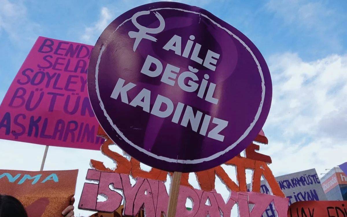 Bu görsel, bir protesto veya kadın hakları eylemi sırasında çekilmiş bir kareyi yansıtmaktadır. Görselde, mor renkli bir pankart dikkat çekmektedir. Pankartta büyük harflerle "AİLE DEĞİL KADINIZ" yazısı yer almakta ve üzerinde kadın sembolü bulunmaktadır. Mor pankart, kadın hareketinin temel renklerinden biri olan morun simgeselliğini taşır ve feminist bir mesaj içerir.