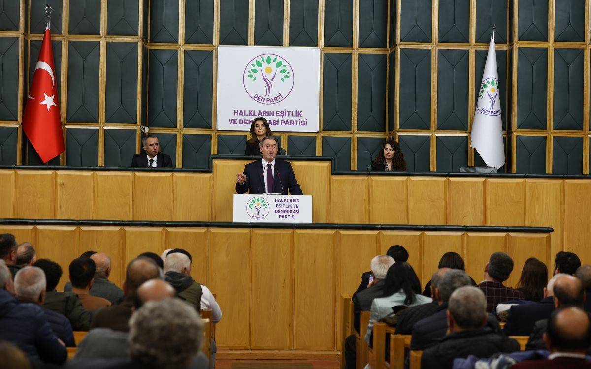 Bu fotoğraf, DEM Parti'nin bir grup toplantısını göstermektedir. Fotoğrafta, kürsüde bir konuşmacı dikkat çekmektedir. Konuşmacının önünde "Halkların Eşitlik ve Demokrasi Partisi" yazılı bir platform yer alıyor ve kürsünün üzerinde DEM Parti'nin logosu ve ismi görülüyor.  Konuşmacının arkasında iki kadın oturuyor, onlar da parti yetkilileri olabilir. Arka planda Türkiye bayrağı ve DEM Parti bayrağı yer almakta. Salonda, ahşap detaylarla çevrili bir dekorasyon görülüyor ve oturma düzeninde çok sayıda kişinin toplantıyı dinlediği anlaşılıyor. Katılımcılar arasında farklı yaş gruplarından bireyler bulunuyor.  Genel olarak, fotoğraf demokratik bir ortamda gerçekleştirilen bir siyasi toplantıyı yansıtıyor ve topluluk önünde yapılan bir konuşmanın dinleyicilere hitap ettiği bir anı kaydediyor.