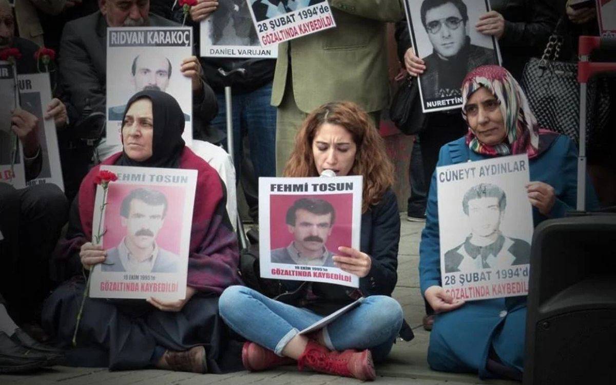 Bu görsel, Cumartesi Anneleri'nin bir eyleminden bir sahneyi gösteriyor. Ön planda, kayıplarının fotoğraflarını tutan üç kadın oturuyor. Soldaki kadın siyah başörtüsü ve kırmızı bir şal giymiş, elinde kırmızı bir karanfil tutuyor. Orta kısımda oturan kadın, mikrofonla konuşurken "Fehmi Tosun - 19 Ekim 1995'te gözaltında kaybedildi" yazılı bir pankart taşıyor. Sağdaki kadın ise başörtülü ve "Cüneyt Aydınlar - 28 Şubat 1994'te gözaltında kaybedildi" yazılı bir pankart tutuyor.  Arka planda, ellerinde diğer kayıpların fotoğraflarını tutan ve ayakta duran kişiler görülüyor. Kayıpların fotoğraflarının altında, kayboldukları tarihlere dair bilgiler yer alıyor. Görsel, Cumartesi Anneleri'nin gözaltında kaybolan yakınları için adalet talebini ve mücadelelerini simgeliyor.