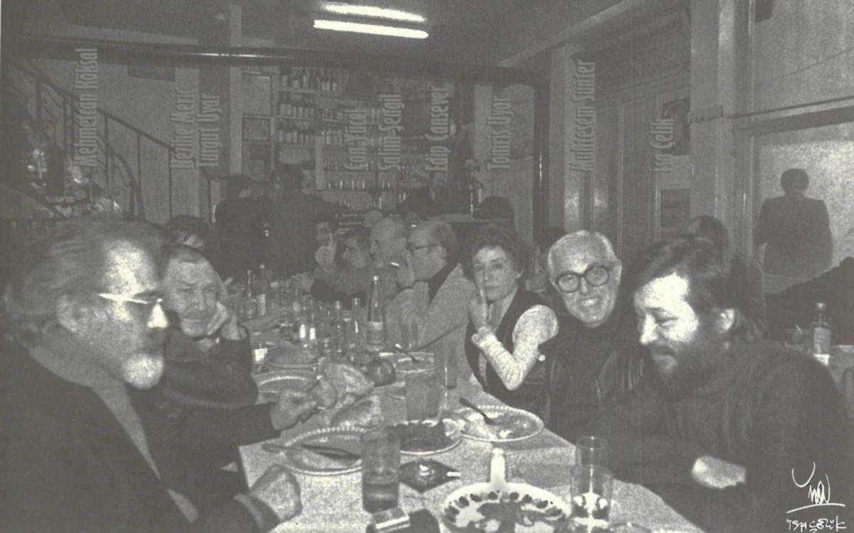 Bir restoran ortamında çekilmiş, samimi bir buluşmaya ait siyah-beyaz bir fotoğraf. Uzun bir masa etrafında oturan insanlar yemeklerini yemekte ve sohbet etmektedir. Masanın üzerinde tabaklar, bardaklar ve yiyecekler görülüyor.