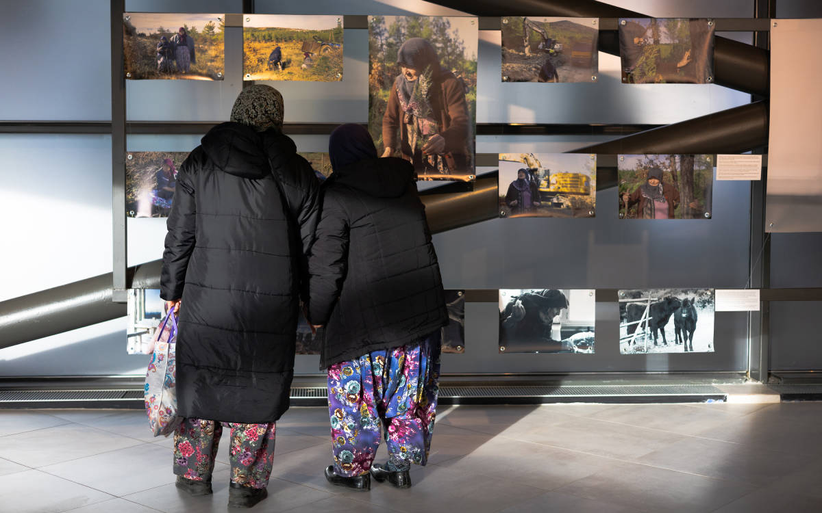 Fotoğrafta, siyah uzun montlar giymiş, başları örtülü ve rengarenk çiçek desenli şalvarlar giymiş iki kadın bir sergiyi inceliyor. Kadınlar sırtları dönük şekilde ayakta duruyor. Önlerinde cam bir pano üzerinde çeşitli fotoğraflar sergilenmiş. Fotoğraflarda doğa, köylü kadınlar ve iş makineleri gibi görüntüler yer alıyor. Arka planda serginin bulunduğu modern bir iç mekân gözükmekte. Kadınlardan biri yanında desenli bir bez çanta taşıyor.