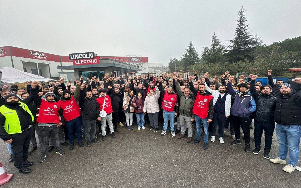 Fotoğrafta, dış mekânda toplanmış büyük bir grup insan görülüyor. İnsanlar topluca kameraya doğru ellerini yumruk yaparak kaldırmışlar; bu, birlik ve dayanışma sembolü olarak yorumlanabilir. Ön tarafta kırmızı yelekler giyen bazı kişiler dikkat çekiyor ve üzerlerinde "GREV GÖZCÜSÜ" yazısı okunabiliyor. Çevrede açık hava koşulları hakim, bulutlu bir gökyüzü var ve grup bir fabrikanın ya da işletmenin önünde toplanmış gibi görünüyor. Arkada “Lincoln Electric” yazılı bir bina tabelası fark ediliyor. Bu, bir grev ya da toplu dayanışma etkinliği gibi duruyor. İnsanların yüz ifadeleri kararlı ve birlik mesajı veriyor.