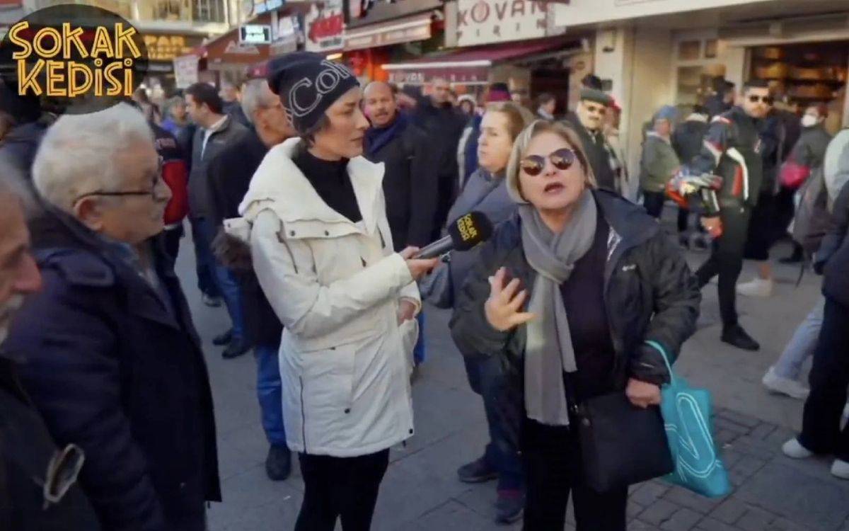 Bir sokak röportajı sırasında çekilen bu görüntüde, kalabalık bir alanda muhabir mikrofon uzatıyor. Mikrofonun üzerinde "Sokak Kedisi" yazıyor. Muhabir, beyaz mont ve siyah bere giymiş. Mikrofonu tutan muhabirin karşısında gözlük takan, siyah montlu ve gri atkılı bir kadın konuşuyor. Çevrede başka insanlar da merakla izliyor veya günlük yaşamlarına devam ediyor. Arka planda dükkan tabelaları ve yoğun bir şehir atmosferi görülüyor.